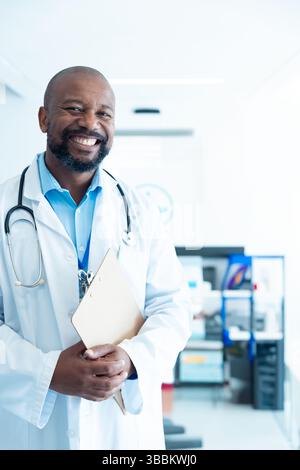 Medico afroamericano senior in piedi in una luminosa stanza d'ospedale che tiene gli appunti e lo stetoscopio Foto Stock