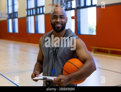 Allenatore di pallacanestro maschile in piedi sul campo da palestra, tenendo gli appunti di pallacanestro e indossando fischio Foto Stock