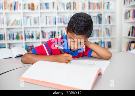 ragazzo birazziale che scrive su un quaderno a griglia al tavolo curvo della biblioteca, usando una matita verde con libri aperti Foto Stock