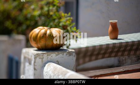 Immagine inclinabile di un ambiente rustico all'aperto con una zucca matura appoggiata su un pilastro di pietra bianca e una caraffa di argilla su una tovaglia a scacchi calda Foto Stock