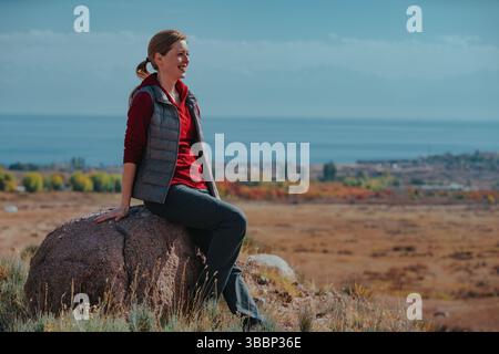 Giovane donna sorridente turistica seduta su pietra sullo sfondo del lago Foto Stock