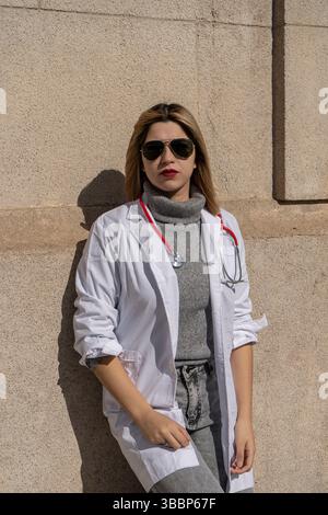 Giovane e sicura medico donna con stetoscopio e occhiali da sole che posano contro una parete Foto Stock