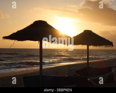 Romantico ambiente con due ombrelloni e lettini di fronte al tramonto sulla spiaggia di Quinto do Lago, sull'Algarve. Foto Stock
