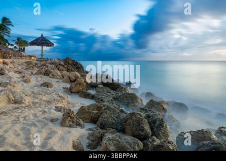 La tranquilla costa al crepuscolo di Aruba presenta onde sfocate dal movimento che si stagliano sulle rocce sotto un cielo blu e nuvoloso, con una palapa da spiaggia. Foto Stock
