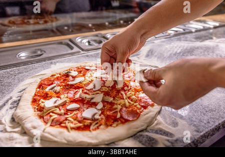 Cuocere in cucina mettendo gli ingredienti sulla pizza. L'uomo prepara la pizza. Cuocere a mano fa una pizza, mette i funghi. Lo chef aggiunge meticolosamente Foto Stock