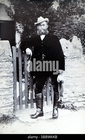 Storia sociale, moda: Un gentiluomo in piedi accanto a un cancello in un ambiente rurale. L'uomo indossa abiti neri, un gilet, stivali al ginocchio e un cappello. Ha una barba grande e tiene una lepre o un coniglio nella mano sinistra. Non attribuito, pubblicato intorno al 1908. Da una collezione mista di cartoline edoardiane. Foto Stock