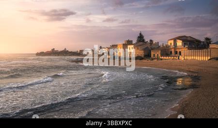 Le onde si infrangono dolcemente contro la spiaggia sabbiosa mentre il sole tramonta dietro le affascinanti case di Punta secca, in Sicilia, offrendo pace, in Italia Foto Stock