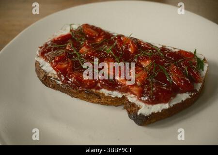 Una fetta di pane tostato ricoperto da uno strato di formaggio spalmabile, marmellata fresca di fragole e erbe di menta tritate Foto Stock