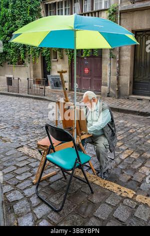Parigi, Francia, 12 luglio 2024: Un vecchio artista con la barba bianca siede su una sedia pieghevole sotto un ombrellone, in una strada acciottolata a Montmartre. Foto Stock