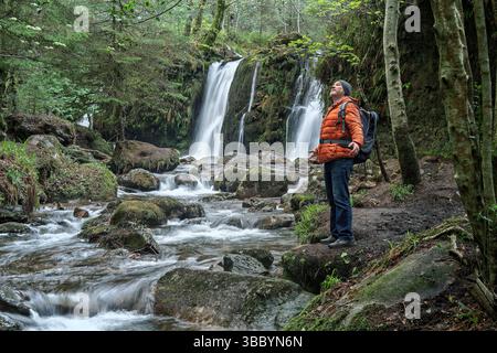 L'uomo abbraccia la bellezza selvaggia di Glenmalure, di fronte alla cascata e al torrente nel profondo della natura selvaggia irlandese. Foto Stock