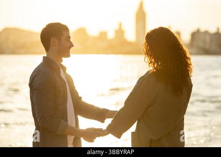 La giovane coppia si trova sulla spiaggia al tramonto, sorridendo e tenendosi per mano mentre la luce dorata soffusa si riflette sull'acqua in una mamma serena e affettuosa Foto Stock