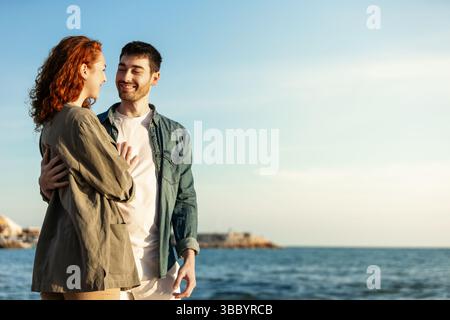La giovane coppia felice si avvicina e sorride calorosamente l'una all'altra, godendo insieme di un momento tranquillo e soleggiato vicino al mare sotto un cielo azzurro. Stile di vita. Foto Stock