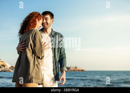 Una giovane coppia felice condivide una risa sincera mentre si abbraccia sulla spiaggia, bagnata dalla luce soffusa del sole con il mare e il cielo blu sullo sfondo. Stile di vita. Foto Stock