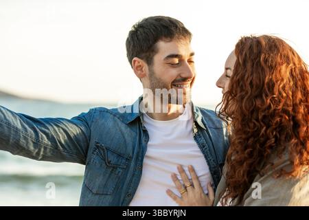 La giovane coppia si trova vicino a una spiaggia, scambiandosi sorrisi e delicati tocchi mentre la luce del sole dorata si riflette sull'oceano sullo sfondo. Stile di vita. VCA Foto Stock