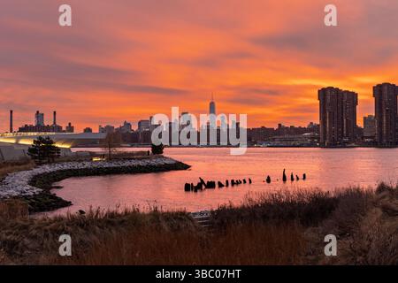 La natura mette in mostra un vibrante tramonto a Long Island City, proiettando un caldo bagliore sullo skyline di New York. L'acqua riflette i colori vivaci degli edifici cittadini Foto Stock