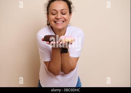 Una donna allegra regala due gustose ciambelle, una con glassa di cioccolato e un'altra rosa smerigliata, su uno sfondo beige, che irradia felicità e felicità Foto Stock