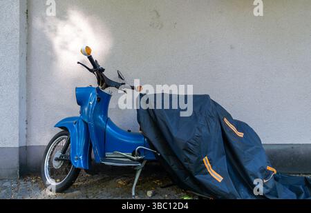 Ciclomotore in stile vintage Sky Blue parzialmente coperto da un telone nero, parcheggiato contro un muro su un marciapiede di Berlino. Foto Stock