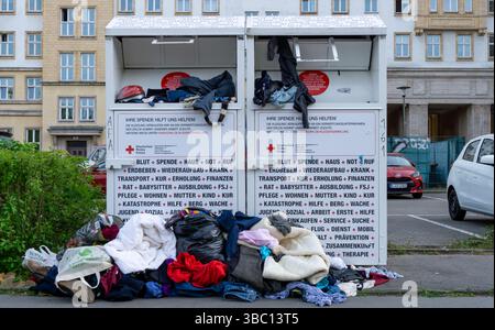 Bidoni per donazioni di abbigliamento traboccanti della Croce Rossa tedesca a Berlino, con indumenti impilati a terra intorno ai contenitori. Foto Stock