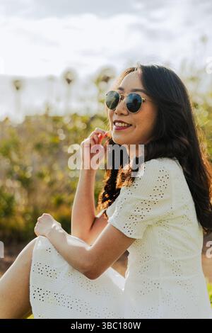 Una donna che indossa occhiali da sole e un vestito bianco è seduta sull'erba. Lei sorride e si sta godendo la vita all'aria aperta Foto Stock