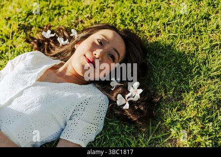 Una donna si sta sdraiando sull'erba con dei fiori nei capelli. Sta sorridendo e guardando la telecamera. Concetto di relax e felicità Foto Stock