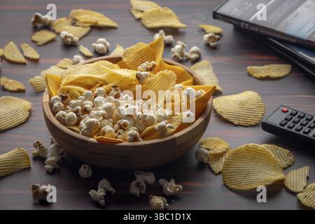 Ciotola in legno con patatine e popcorn sparsi sul tavolo con telecomando e DVD Foto Stock