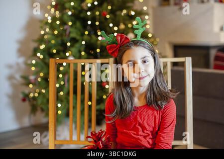 La ragazza della scuola elementare seduta con un dono sullo sfondo dell'albero di Natale. La ragazza vestita con un abito rosso ha un cerchio sulla sua testa con de Foto Stock