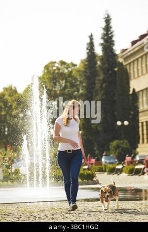 Una giovane donna che cammina con un cane da aquila su un guinzaglio vicino alla fontana nel parco Foto Stock