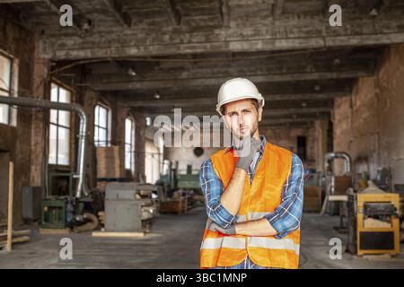 Il giovane perplesso con un casco protettivo e un giubbotto arancione in piedi nella vecchia fabbrica durante l'installazione dell'attrezzatura. Lavoratore premuroso in piedi Foto Stock