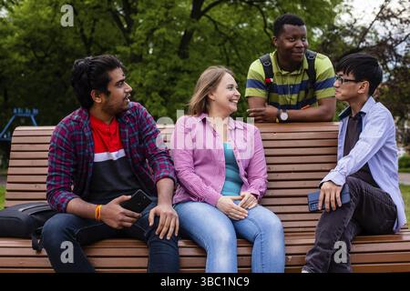 Azienda variegata di giovani uomini e donne su panca di legno su sfondo verde alberi. Amici che trascorrono del tempo a chiacchierare all'aperto. Conc. Amicizia multirazziale Foto Stock