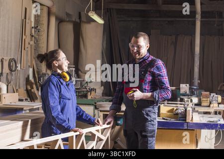 Ritratto di uomo e donna, falegnami in abbigliamento da lavoro, che fa arredamento in legno per letto. Team creativo di marito e moglie che lavorano insieme in mobili Foto Stock