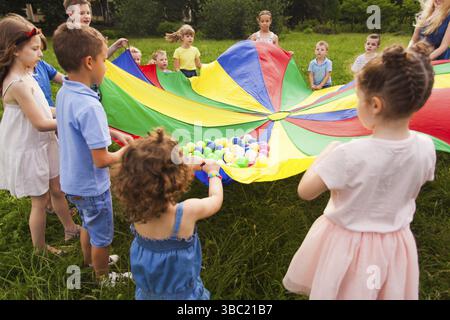 Vista ravvicinata dei bambini di età diversa che tengono stretto il paracadute colorato con delle palline su di esso. Bambini che rotolano sopra le palle piccole sul paracadute, trasmettendo la massa Foto Stock