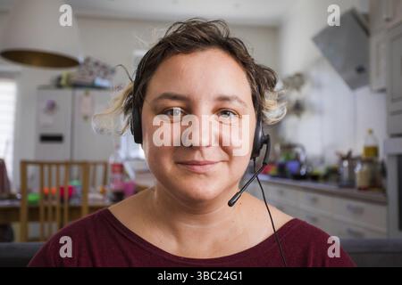 Ritratto di giovane donna attraente in cuffie durante la sua conference call. Insegnante che fornisce lezioni online durante la quarantena. Effetto di ripresa con la webcam Foto Stock