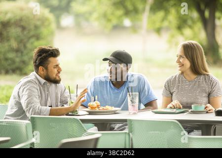 Diversi studenti mangiano insieme. Gruppo di giovani allegri seduti sulla terrazza. Pranzo all'aperto nel weekend estivo Foto Stock