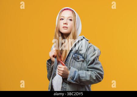 Elegante giovane donna in felpa bianca con cappuccio sulla testa e giacca in jeans, che si posa con un look audace stretto alla fotocamera. I giovani non hanno limiti con Foto Stock