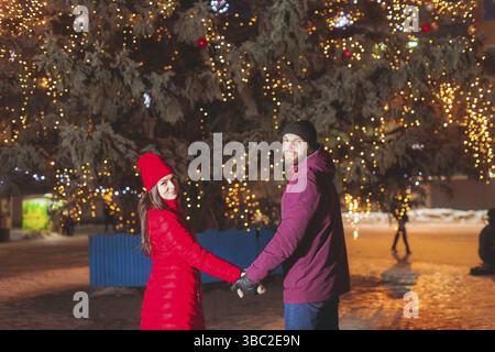 Ritratto di giovane coppia attraente posa vicino città albero di Natale decorato con luci. Sposi che viaggiano durante le vacanze invernali, indossando un caldo stile Foto Stock