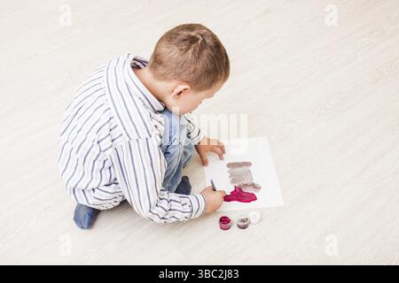 Ragazzo è dipinto con un pennello e acquerelli su carta sul pavimento Foto Stock
