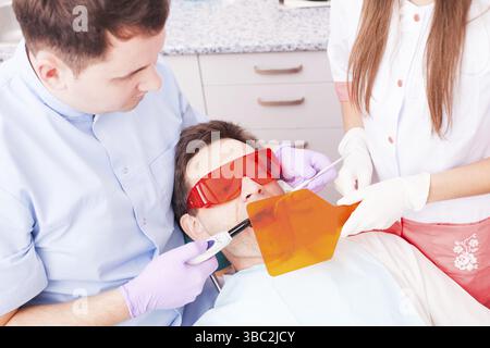 Il paziente giace in speciali occhiali di protezione contro radiazioni laser mentre il dentista lavora con dentale luce di indurimento Foto Stock