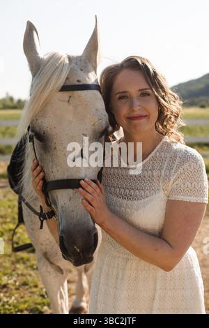 Ritratto di una giovane donna in un abito bianco che abbraccia il cavallo al ranch Foto Stock