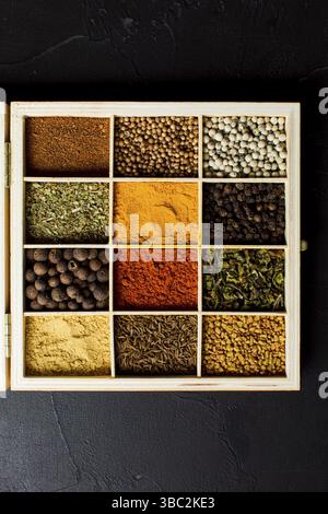 Stoccaggio in legno per le spezie. Scatola divisa in celle, riempita con diverse erbe secche e spezie. Condimenti colorati e diversi, vista dall'alto sul dorso nero Foto Stock