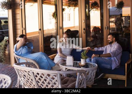 Tre amici felici stanno parlando e ridendo mentre si siedono in una terrazza del coffee shop. Due donne e un uomo comunicano spensierati Foto Stock