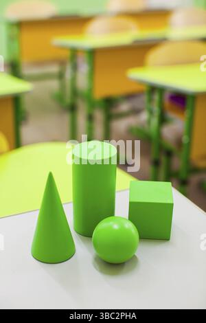 Chiudere le varie figure geometriche sulla superficie bianca. Aula con scrivanie gialle sullo sfondo. Forme geometriche verdi per la lezione scolastica Foto Stock