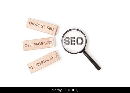 "Tre blocchi di legno etichettati "SEO on-page", "SEO off-page", "SEO tecnico" accanto a una lente di ingrandimento che concentra la parola "SEO" su sfondo bianco. Foto Stock