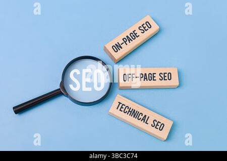 "Tre blocchi di legno etichettati "SEO on-page", "SEO off-page", "SEO tecnico" accanto a una lente di ingrandimento che concentra la parola "SEO" su sfondo blu. Foto Stock