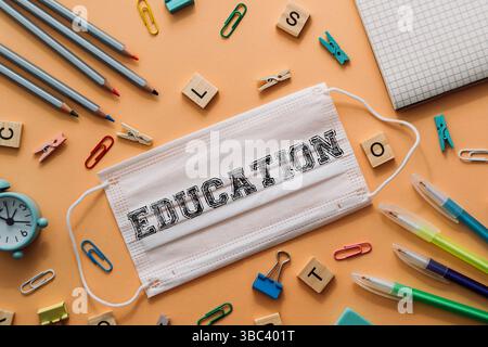 Maschera bianca con la parola "educazione" scritta sopra. La maschera si trova sulla parte superiore di un tavolo con varie forniture per ufficio, come penne, matite e graffette Foto Stock