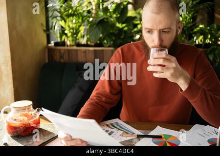 Uomo barbuto in maglione caldo con un conto e altri documenti che bevono tè, concentrandosi sulla gestione delle sue finanze seduti in un caffè verde. Foto Stock