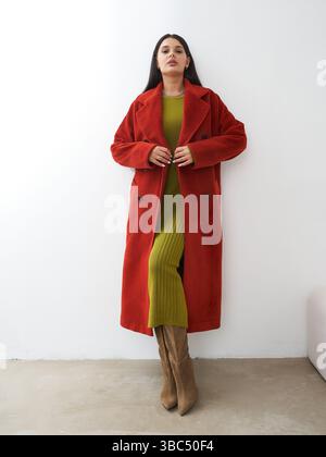 Una donna dai lunghi capelli scuri è sicura, indossa un cappotto rosso vivace su un abito verde testurizzato e stivali al ginocchio marrone. Si piega leggermente Foto Stock