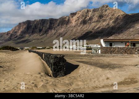 Spiaggia sabbiosa a Famara su Lanzarote con una facciata in pietra e case in collina. Un muro di pietra separa la sabbia del Sahara soffiata da una collezione di edifici bianchi. Urbanizacion Famara, Urbanización Famara, Canarias, Spagna Foto Stock