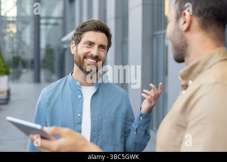 Due uomini in un ambiente di lavoro sono impegnati in una conversazione, l'uomo con la barba sembra felice. Uno sta tenendo un tablet. Foto Stock