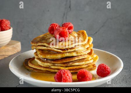 primo piano di una pila di frittelle di banana dorate su un piatto bianco, ricoperte di lamponi freschi e condite con sciroppo Foto Stock
