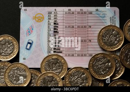 Patente di guida del Regno Unito che mostra le categorie di veicoli e le date di validità circondate da monete inglesi da una sterlina sulla superficie nera, simboleggiando la guida legale e la fi Foto Stock
